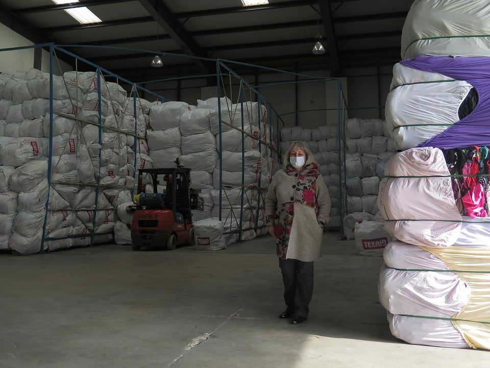 Eine Frau steht in einer Lagerhalle neben einem Turm aus gebrauchten Textilien.