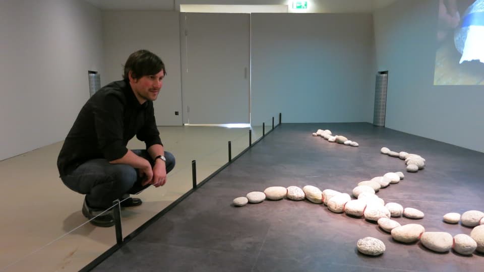 Florian Gredig kniet vor einer Instalation und betrachtet sie: helle Steine auf einer grauen Bodenfläche, angeordnet in Form einer Schlaufe.