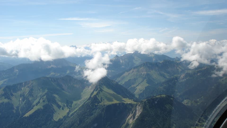 Flugaufnahme auf Berglandschaft: An den Gipfeln hat es kleine Wolken, der Himmel ist sonst blau. 