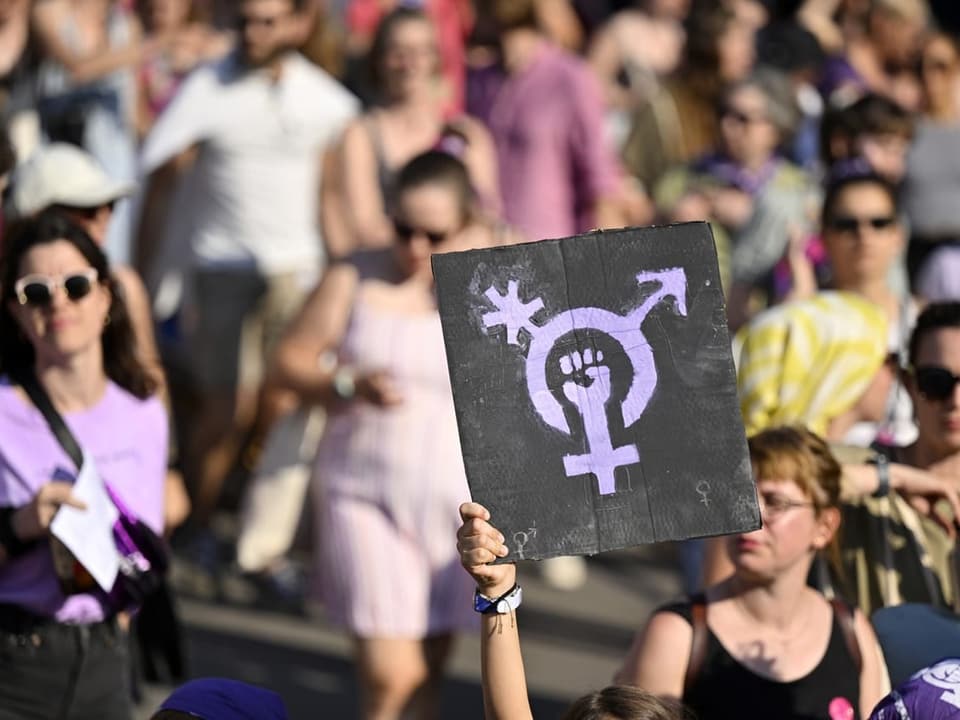 Frau an Frauenstreikdemonstration. Sie hält ein Plakat mit dem Zeichen für Geschlechtervielfalt.