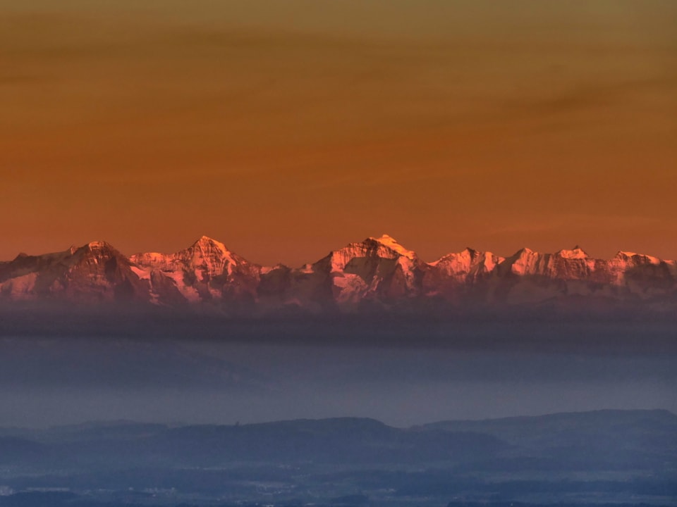 Oranger Himmel, Berge mit verschneiten Spitzen, unten Dunstschicht.
