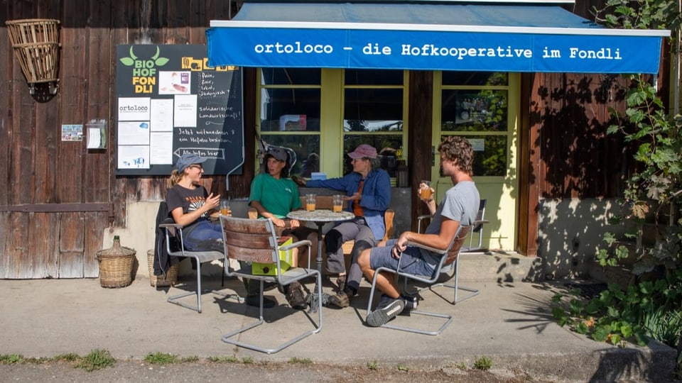 Vier Menschen sitzen vor einer alten Hofwand um einen Tisch, auf Sonnenschutz steht «ortoloco».