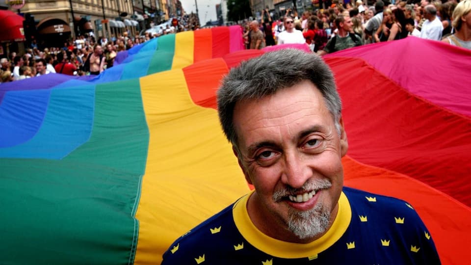 Ein Mann vor einer riesigen Regenbogenfahne an einer Parade.