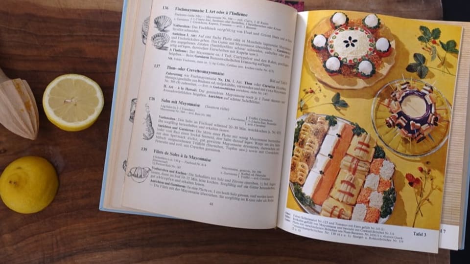 Ein aufgeschlagenes Kochbuch, rechts Farbillustration von bunten Platten, links eine Zitrone auf dem Holzbrett