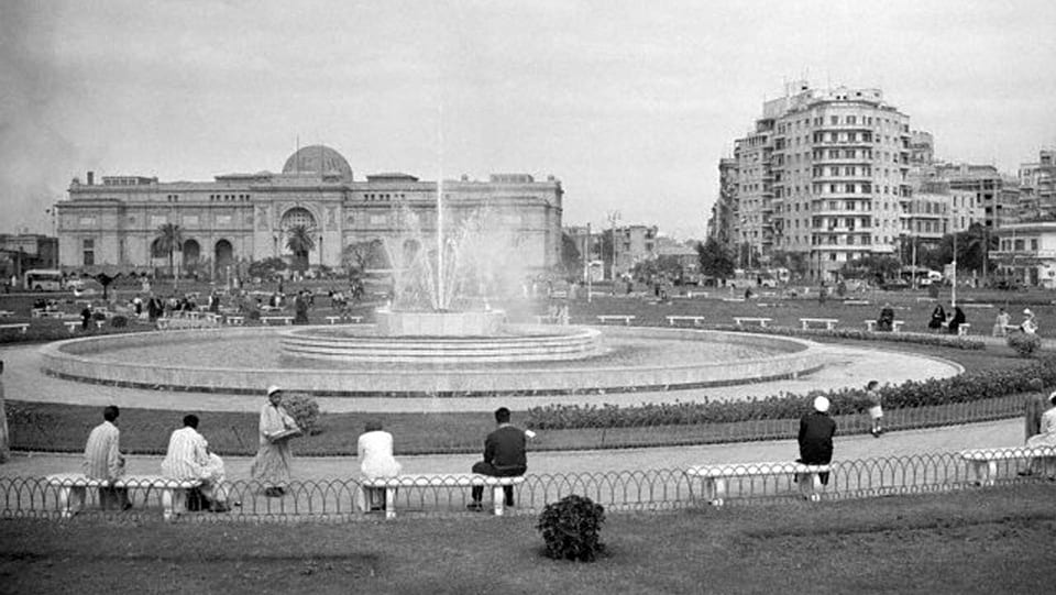 Schwarzweissfoto eines grossen Platzes mit rundem Springbrunnen, im Hintergrund ein grosses, palastartiges Gebäude.