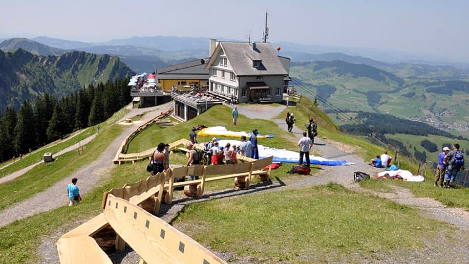 Blick über Bergkrete auf Berggasthaus. Im Vordergrund eine lange Holzbank. 