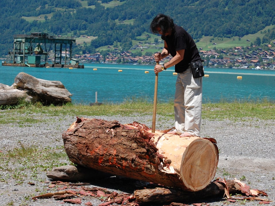Naruo Nishimura bearbeitet einen Baumstamm mit einem Schäleisen am Seeufer.
