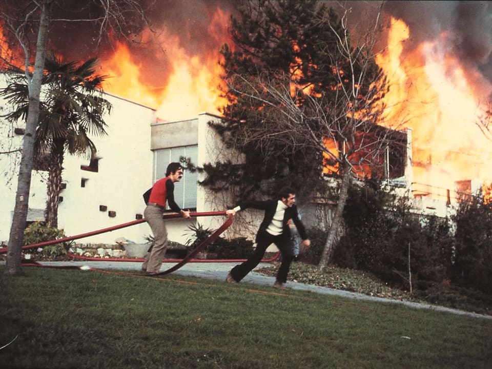 Zwei Männer, einer am Feuerschlauch. Im Hinterrund brennt ein Gebäude.