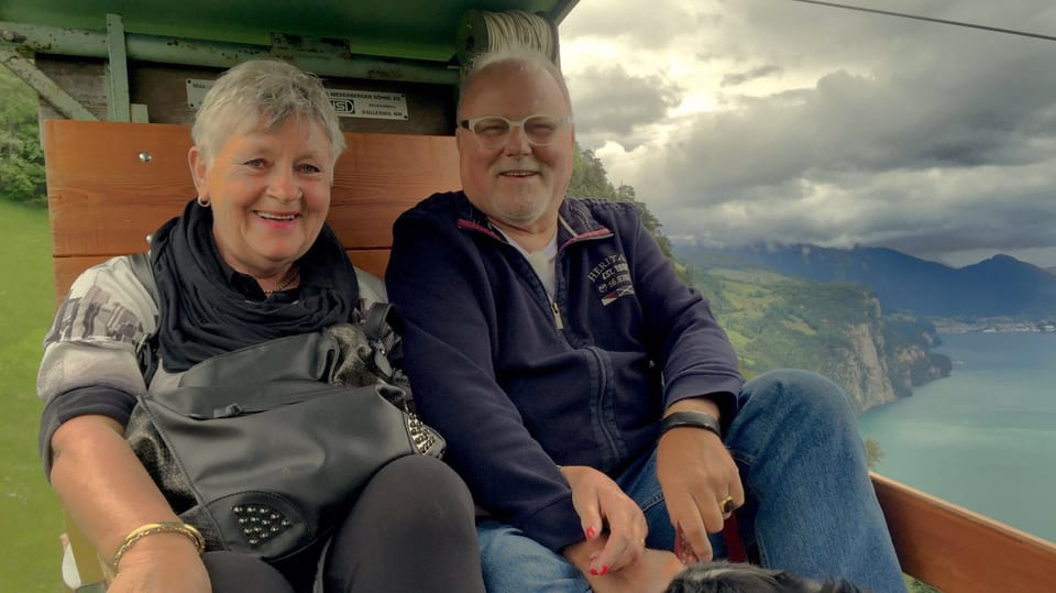 Ein älteres Ehepaar sitzt in einer offenen Seilbahn, beide lachen. Neben der Bahn sieht man 800 Meter in die Tiefe zum Vierwaldstättersee.