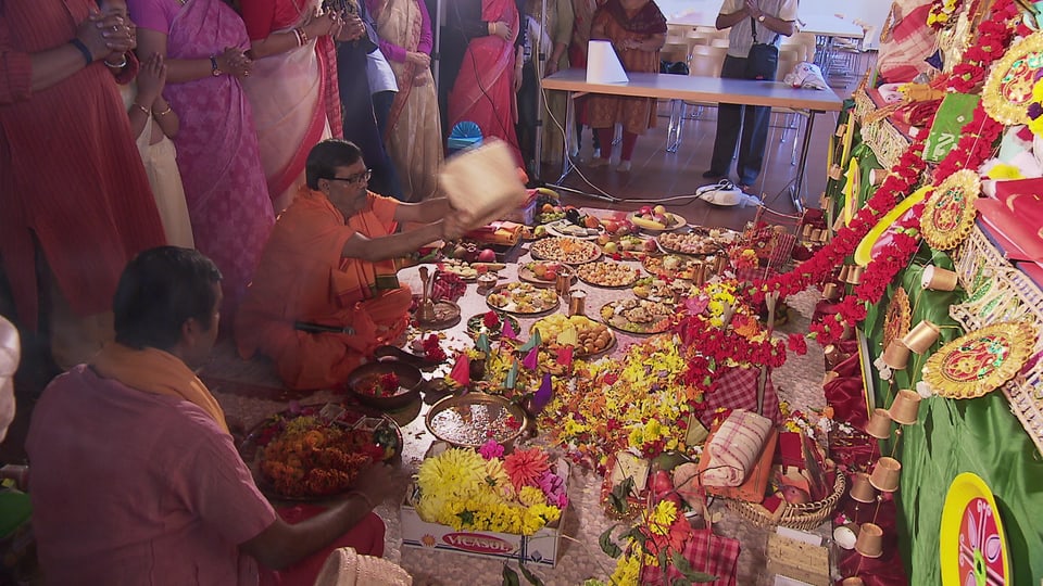 Auf dem Boden sind Speisen vor einem Altar ausgebreitet. Menschen stehen und sitzen davor.