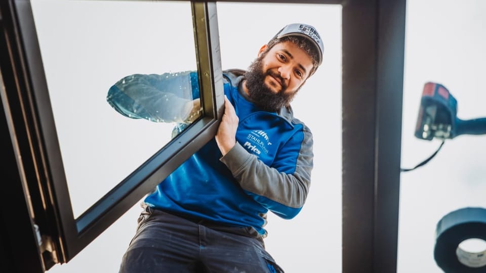 Mann mit dunklem Bart und Mütze in blauer Arbeitsjacke schaut von oben runter, hält Fenster.