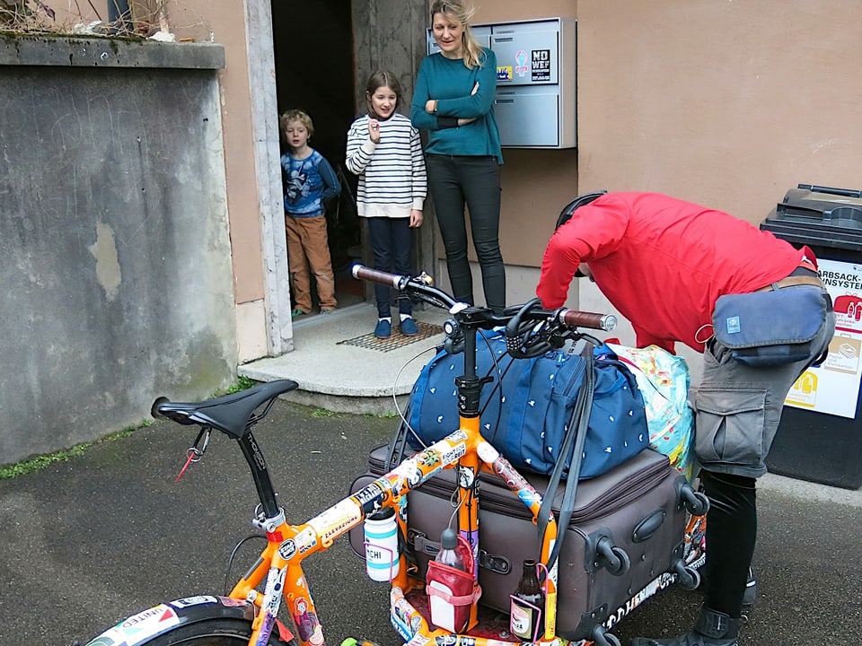 Frau und zwei Kinder stehen bei ihrer Haustür und schauen einem Velokurier zu, der ihr Gepäck auf sein Cargovelo lädt.