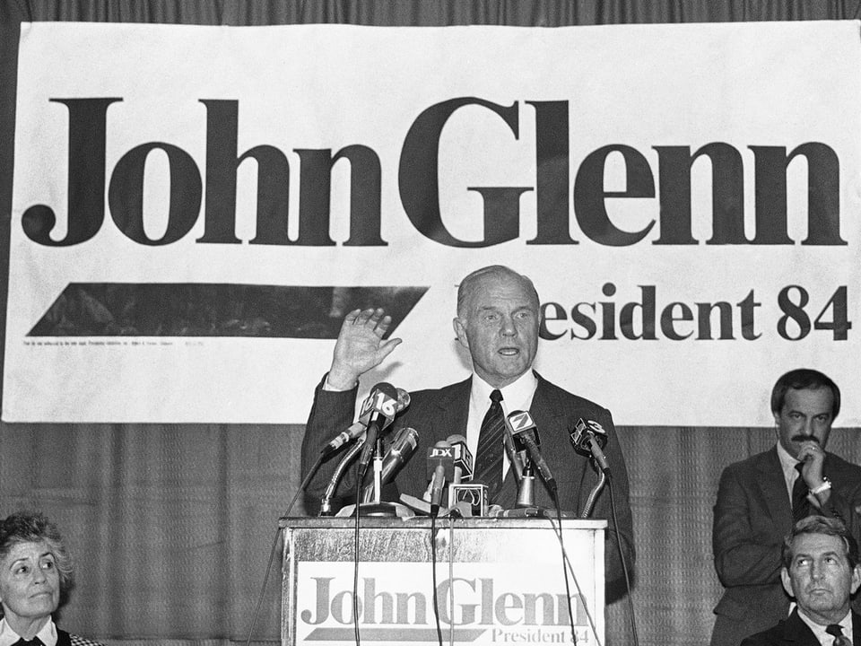 Glenn steht vor einem Rednerpult, dahinter Plakat mit Aufschrift John Glenn President 84