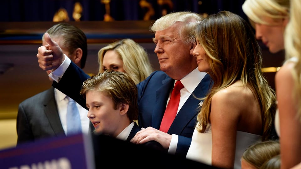 Donald Trump steht mit Frau und Sohn und weiterer Entourage am Rednerpult und reckt den rechten Daumen in die Höhe.