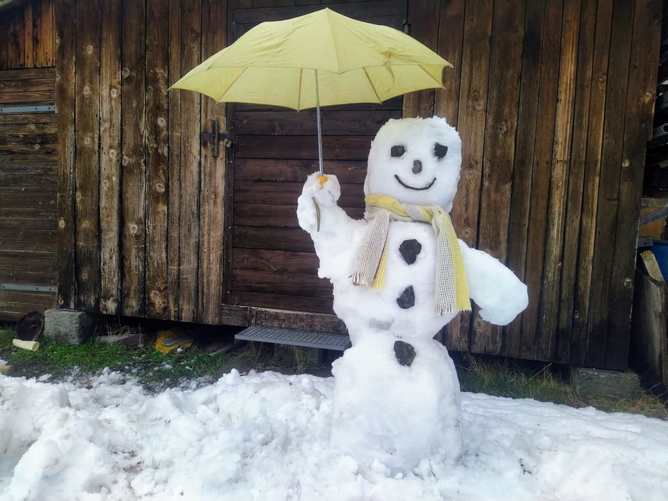 Ein Schneemann vor einem Holzhaus. Er hält einen gelben Regenschirm in der Hand.
