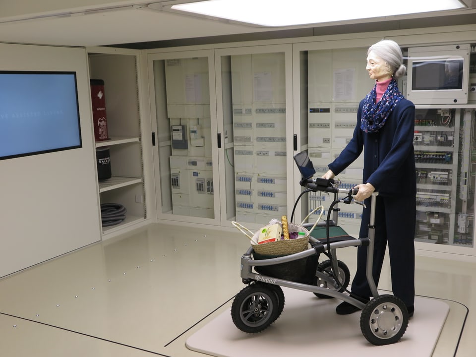 In einem Raum mit vielen Apparaten steht eine lebensgrosse Puppe die sich auf einen Rollator stützt. 