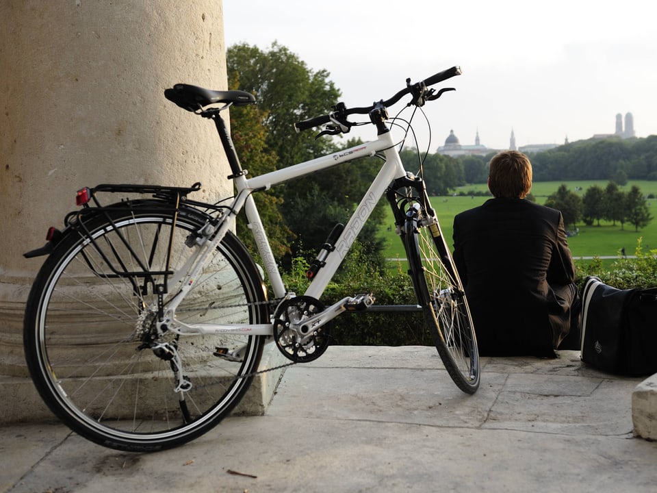 Fahrrad lehnt an einer Säule. Davor sitzt ein Mann, der auf einen Park schaut.