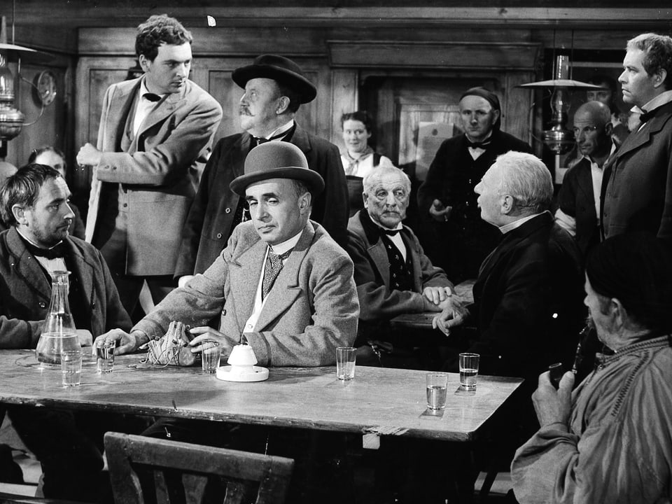 Mehrere Männer in einer Kneipe. Auf dem Tisch im Vordergrund stehen Sechs Gläser. Die Männer blicken sich alle mistrauisch an.