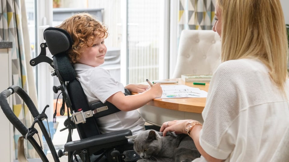 Ein kleiner Junge im Rollstuhl mit der Krankheit Muskeldystrophie Duchenne sieht lächelnd seine Mutter an.