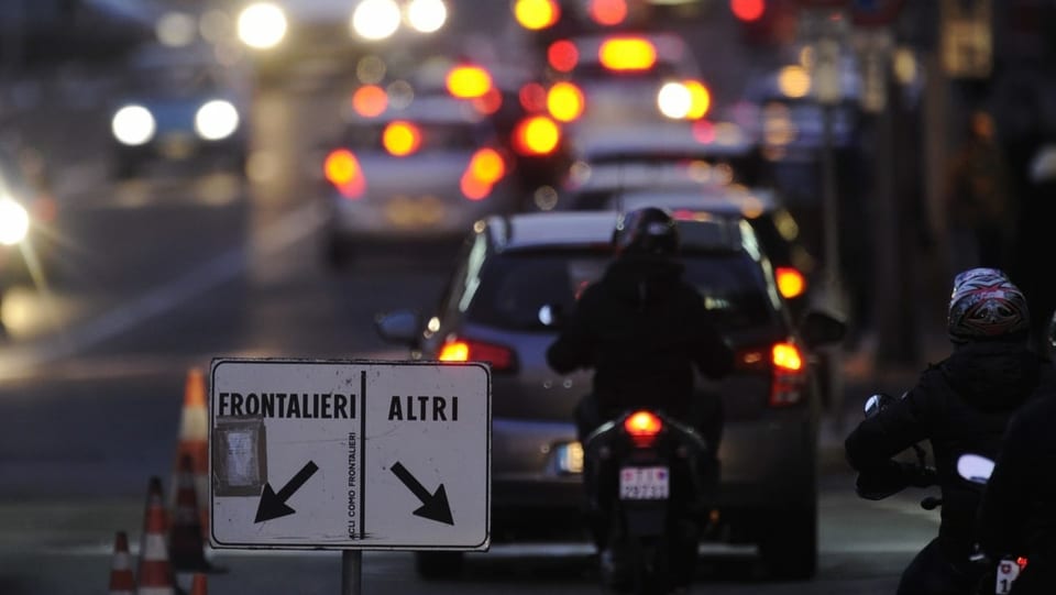 An der Grenze. Autos und Motos stehen Schlange. Auf einem Schild steht frontalieri und altri. Es ist abends.