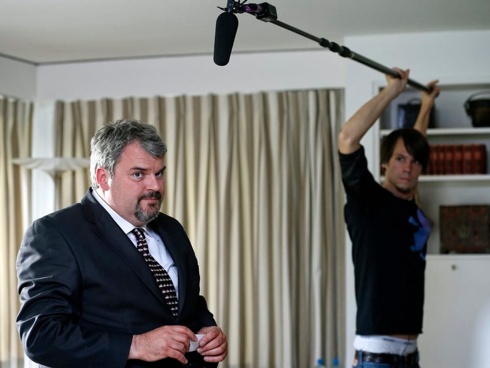 Müller steht in einem Wohnzimmer, im Hintergrund der Tontechniker mit einem Stabmikrofon.