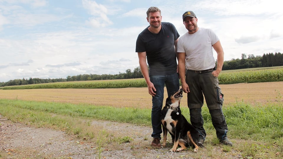 Zwei Männer und ein Hund stehen auf einem Feld.
