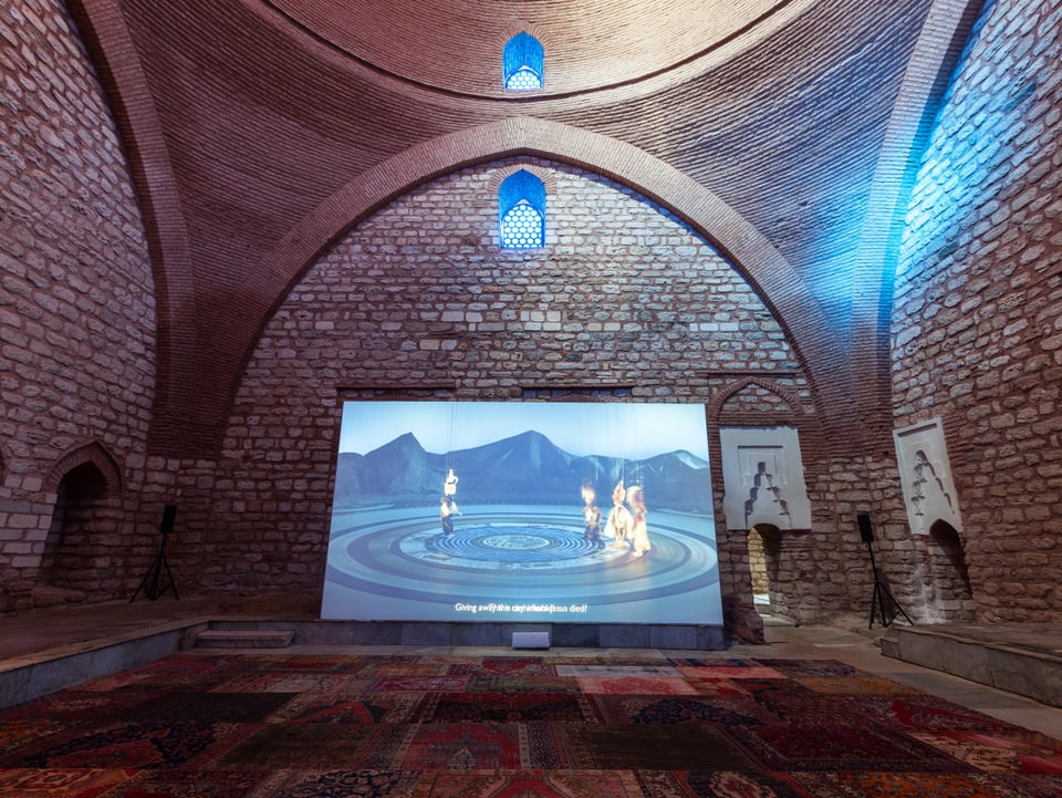 In einem Innenraum einer islamischen Architektur befindet ist eine Videoprojektion zu sehen. Der Boden ist mit zahlreichen angeschnittenen Teppichen ausgelegt.