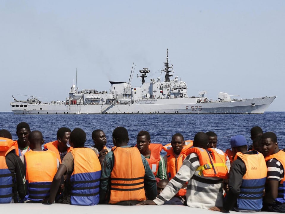 Flüchtlinge auf einem Boot vor einem Schiff der italienischen Marine.
