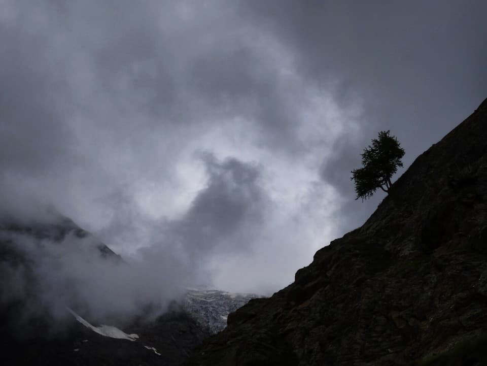 Dunkel graue, hellgraue und auch weisse Wolkenfetzen dominieren das Bild. Im unteren Teil sind dabei dunkel Felsen auszumachen. Am rechten Hang zeichnet sich vor den Wolken die Silhoutte eines Baumes ab.