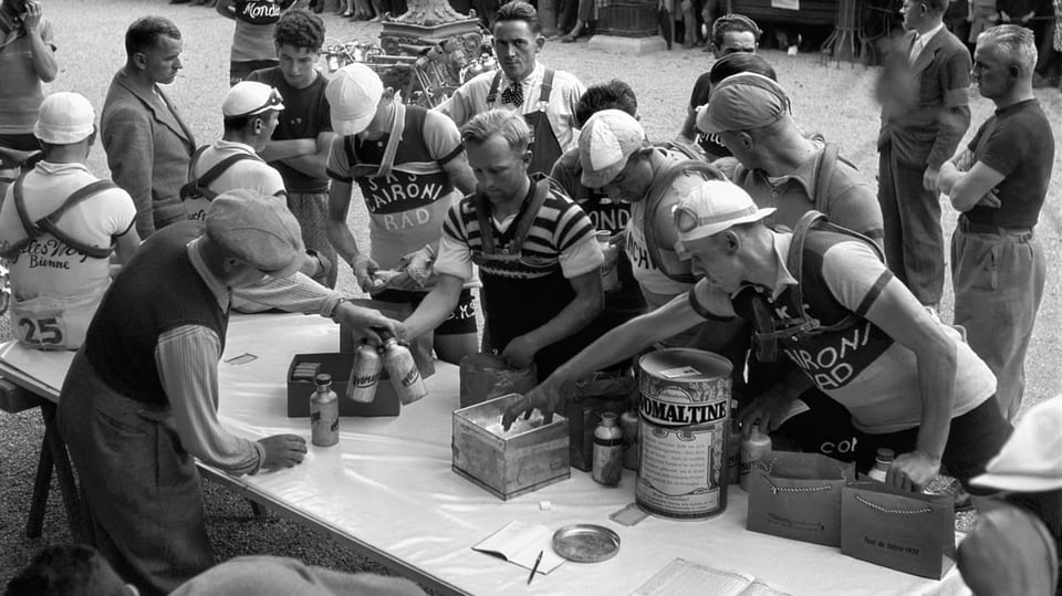 Fahrer der Tour de Suisse 1938 holen sich an einem Verpflegungsstand neue Ovomaltine-Bidons, Ovomaltine-Riegel und Traubenzucker.