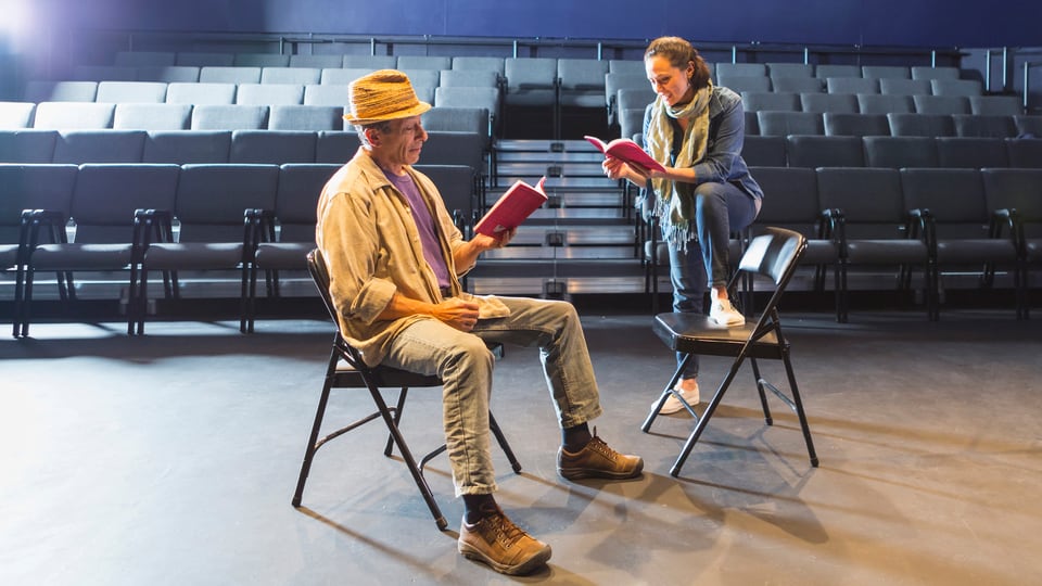Ein Mann und eine Frau sitzen im leeren Theatersaal und halten ein Heft ind en Händen.