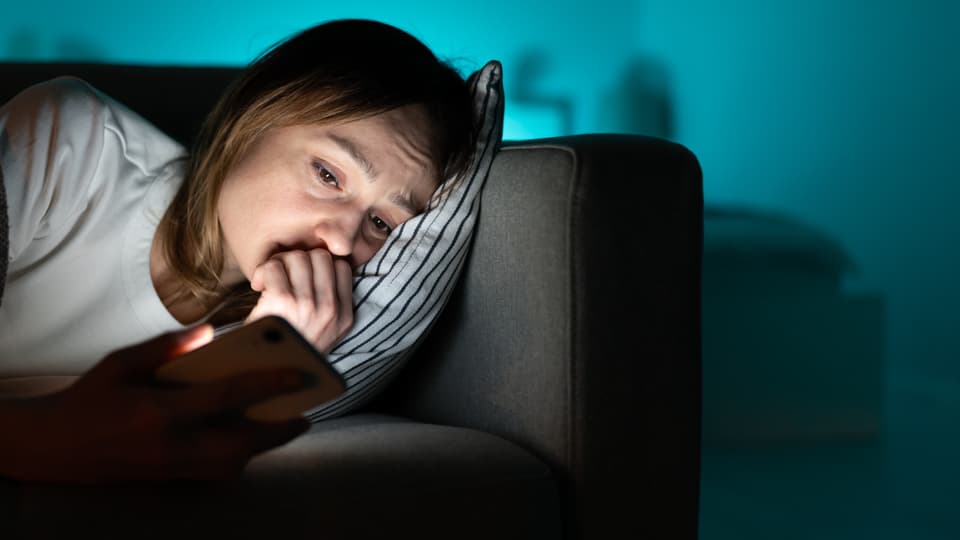 Ein Mädchen oder eine junge Frau liegt im Dunkeln auf der Couch und schaut auf ihr Smartphone. Sie sieht traurig aus.