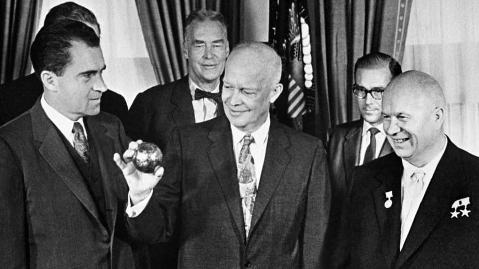 Präsident Eisenhower hält eine Metallkugel. Rechts steht Nikita Chruschtschow, links Vizepräsident Richard Nixon.