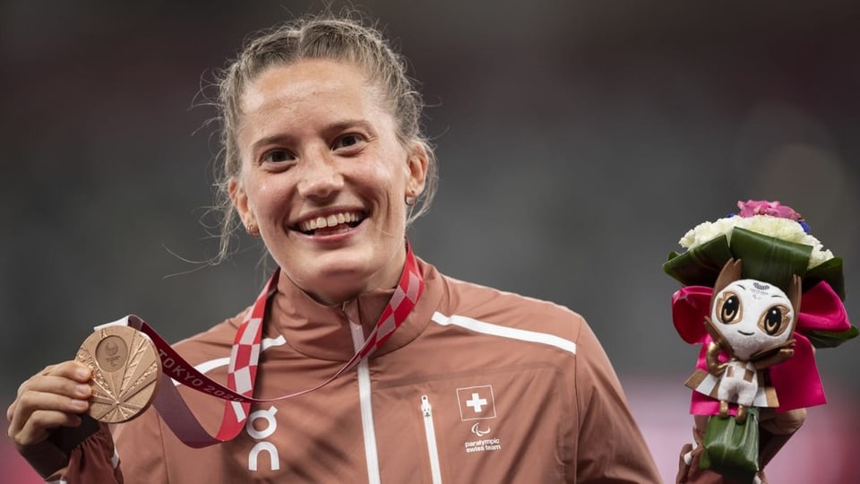 Blonde, junge Frau in rotem Trainer lächelt und hält eine bronzene Medaille vor ihre Brust