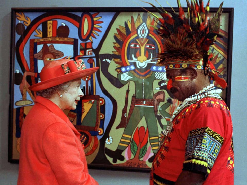 In der linken Bildhälfte sieht man die Queen  mit einem roten Mantel und einem roten Hut. Recht davon steht ein Künstler aus Papua-Neuguinea mit einem traditionellen Hemd und einem Federhut – ebenfalls in rot. Im Hintergrund sieht man ein Bild des Künstlers.
