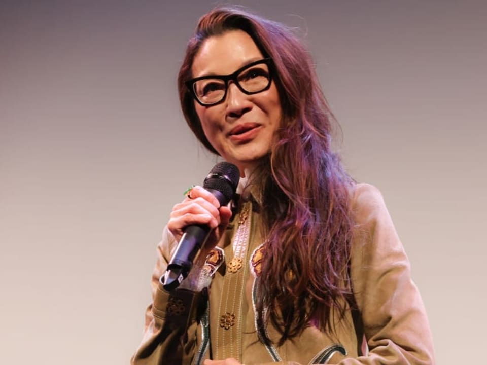 Frau mittleren Alters mit langem dunkeln Haar und grosser schwarzer Brille hält Mikrofon in der Hand und lächelt.