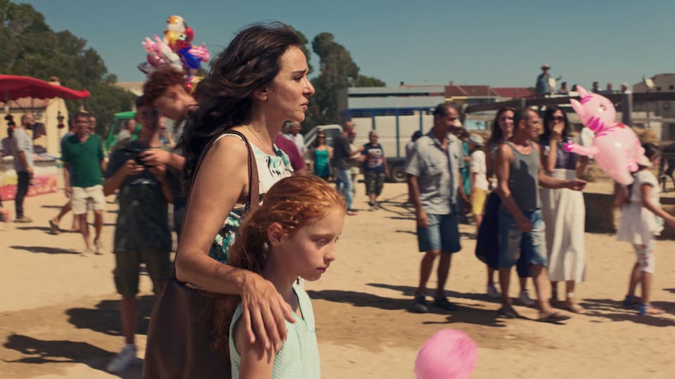 Auf einem Rummelplatz, im Hintergrund Menschen mit Ballons. Im Vordergrund eine Frau, die die Hand um ein Mädchen gelegt hat.