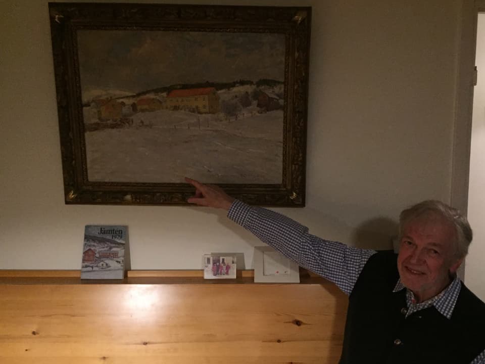 Heinz Erlbacher sitzt auf einem Holzbank und zeigt auf ein Gemälde.