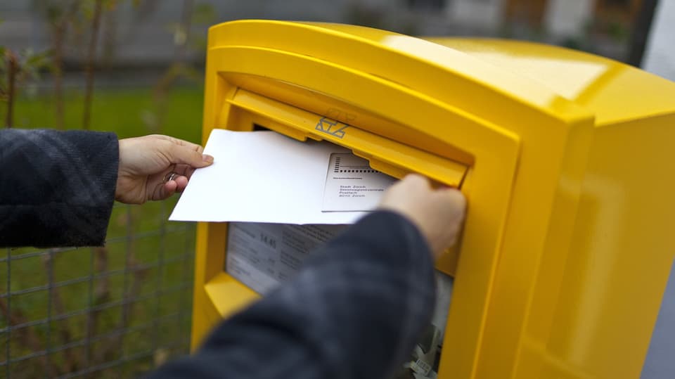 Hände stecken ein Couvert in einen Briefkasten.