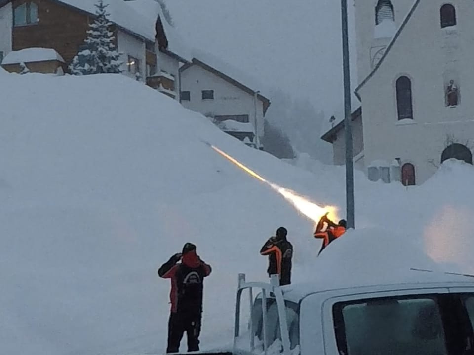 Zwischen Häusern liegt sehr viel Schnee. Drei Männer hantieren im Halbdunklen mit Lampen und sind am sprengen. 
