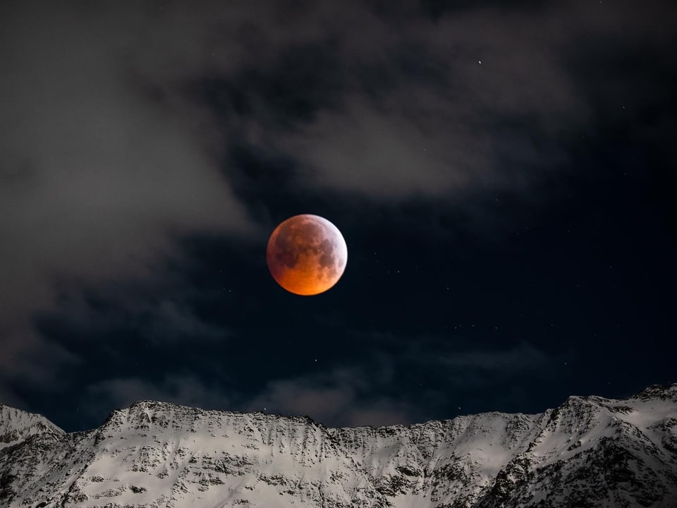 Der rötlich leuchtende Vollmond - während der totalen Mondfinsternis - steht über verschneiten Bergen .