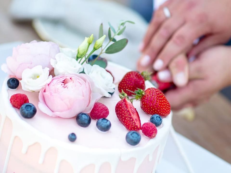 Ein Brautpaar schneidet den Kuchen gemeinsam an. Der ist dekoriert mit Blumen, Erd- und Blaubeeren.
