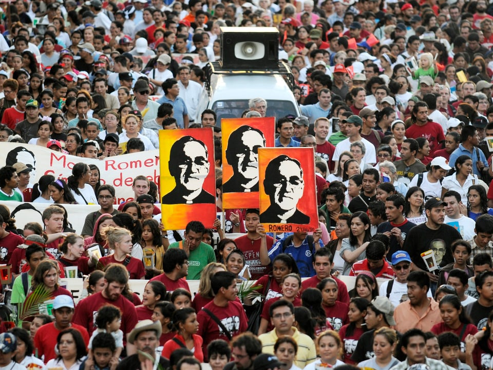 Menschen demonstrieren und halten Schilder mit dem Gesicht von Romero in die Höhe 