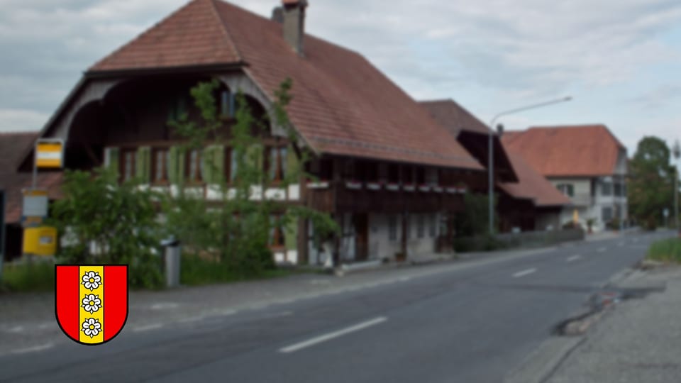 Eine Strasse mit einem Bauernhaus, davor das neue Wappen der Gemeinde.