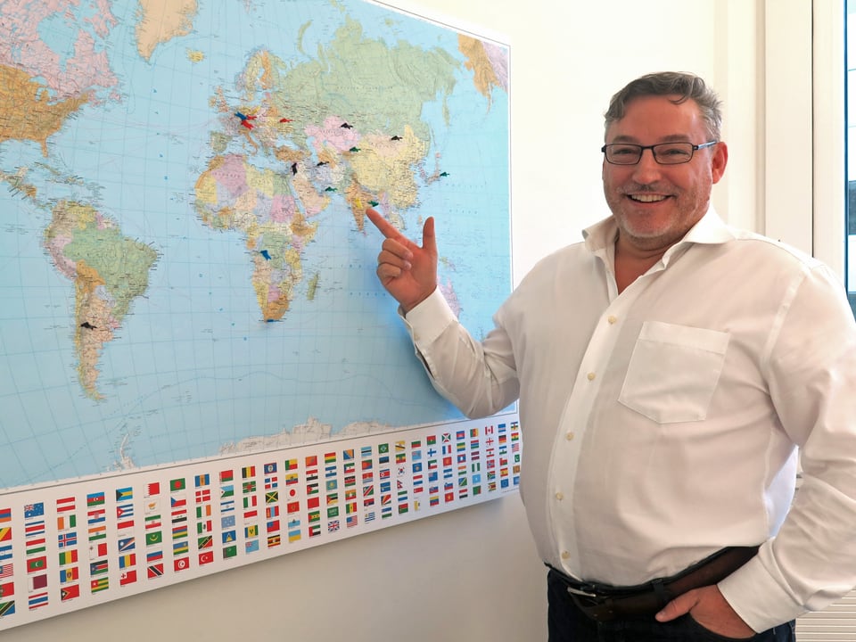Mann mit weissem Hemd vor einer Weltkarte, auf der an verschiedenen Orten Nadeln stecken