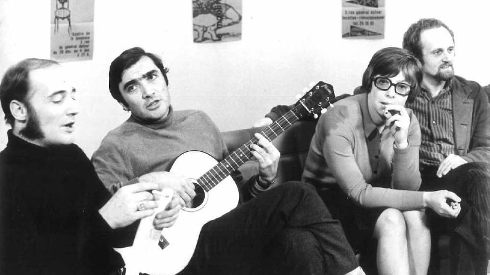Drei Männer und eine Frau sitzen nebeneinander auf einem Sofa. Einer hält eine Gitarre in der Hand.