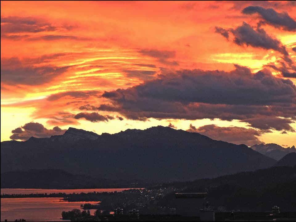 Eine Wolkendecke am Himmel leuchtet orange-rot. Unten ist eine Landschaft mit Ufer, See und Bergen zu sehen, alles noch im Dunkeln. 