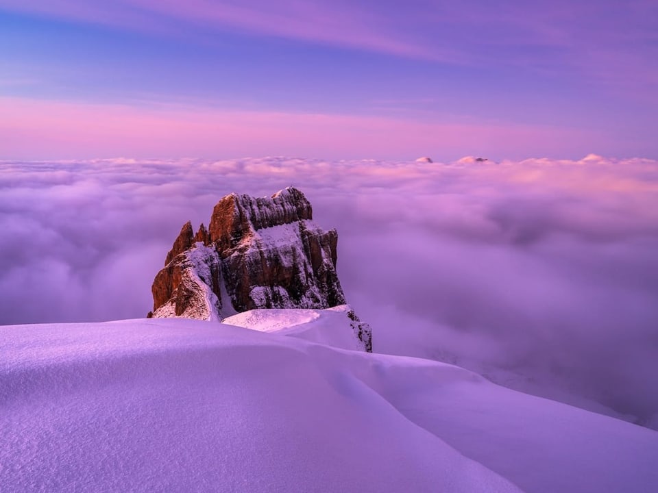 Bergfoto mit Neuschnee und Felsen, im Tal Nebel, alles in violettem Licht der Dämmerung. 