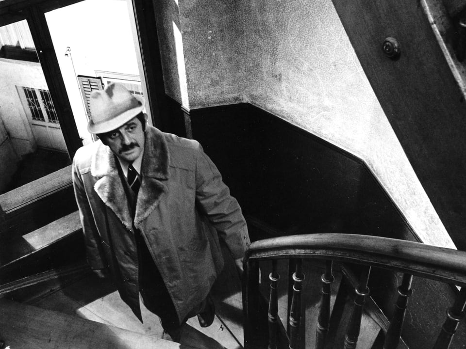 Ein Mann mit Mantel und Hut steht in einem Treppenhaus und blickt nach oben.