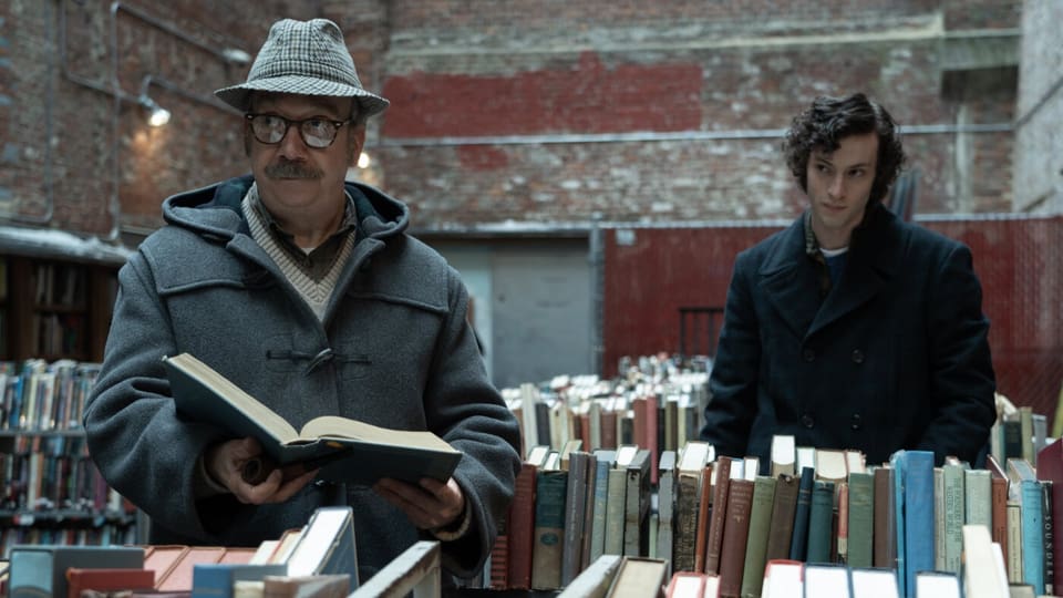 Ein älterer weisser Mann mit Hut und Schnauz blättert in einem Buch, während ein junger weisser Mann ihn beobachtet